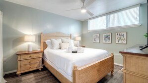 Un dormitorio con cama de madera, dos mesitas de noche con lámparas, ventilador de techo y ventana con persianas.