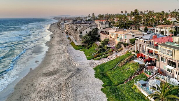 Una zona residencial costera con propiedades frente a la playa, incluyendo edificios de varios pisos con balcones y un paseo marítimo.