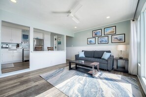 Ein modernes Wohnzimmer mit einem grauen Sofa, einem Couchtisch und einem Teppich. Durch einen offenen Durchgang ist eine Küche zu sehen.