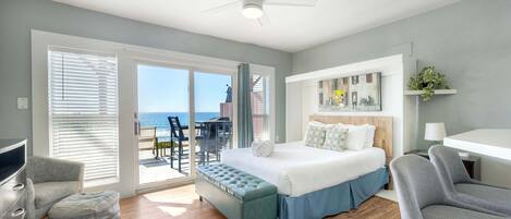 A modern hotel room with a bed, a desk, a chair, a bench, and a view of the ocean through the sliding glass door.