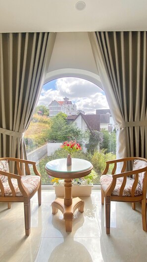 A room with a large window, two chairs, a small table with a vase of flowers, and a view of buildings and greenery outside. - Loc Phu Villa - Hoanglee (Lam Vien - Da Lat)