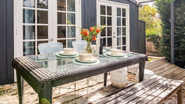 Ein Tisch mit Glasplatte, weißem Geschirr und einem Blumenstrauß, der auf einem Korbtisch steht, in einer überdachten Außenbereich mit Holzterrasse.