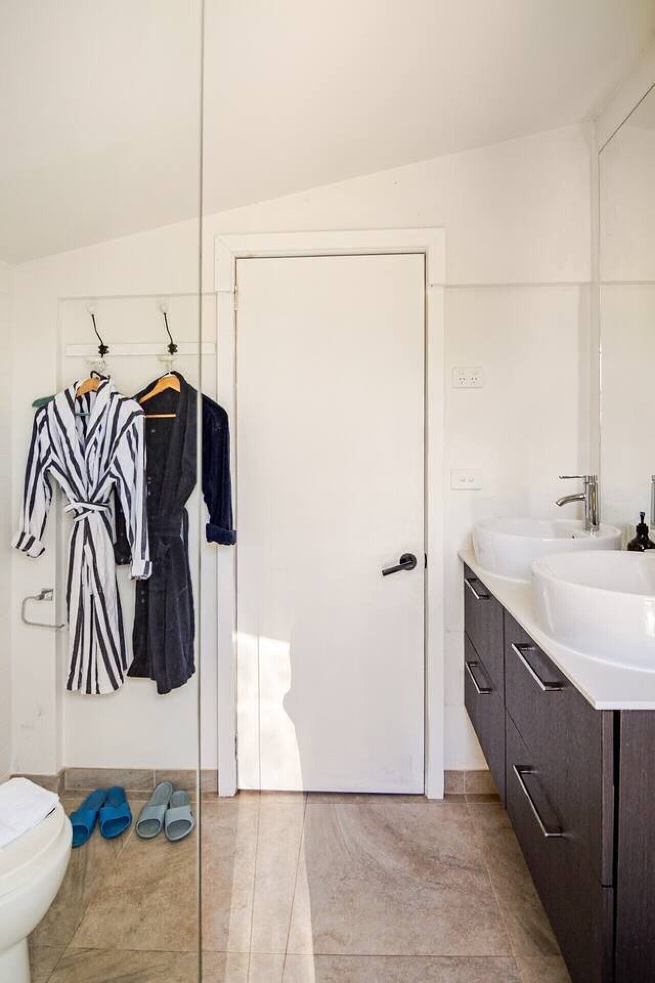 A bathroom with a white door, two sinks, dark cabinets, and a towel rack with robes.
