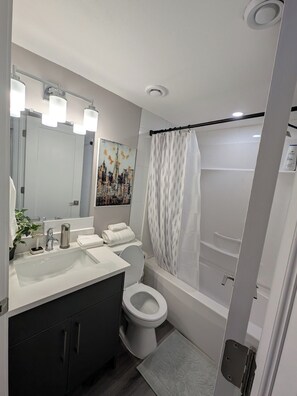 A modern bathroom with a white sink, a toilet, and a bathtub. There is a shower curtain with a leaf pattern, a mirror above the sink, and a plant on the left side. - A serene and stylish one-bedroom suite designed to make you feel at home (Saskatoon)