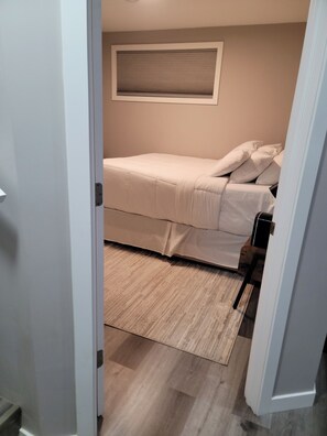 A bedroom with a bed, a window with blinds, and a wooden floor. - A serene and stylish one-bedroom suite designed to make you feel at home (Saskatoon)