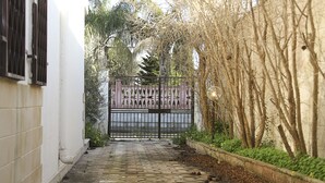 Un étroit sentier mène à une zone délimitée par une grille, avec un bâtiment blanc d’un côté et une clôture bordée d’arbres de l’autre.