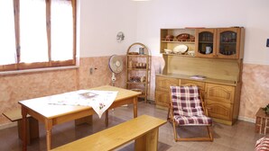 Un coin repas avec une table en bois, un banc et une chaise. On y trouve un ventilateur, une étagère avec de la vaisselle et une fenêtre avec des stores.