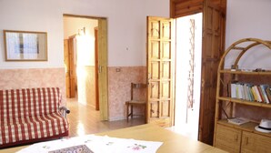 Une pièce avec un canapé à carreaux, une table en bois recouverte d’une nappe à motifs floraux, une bibliothèque et une porte donnant sur une autre pièce.