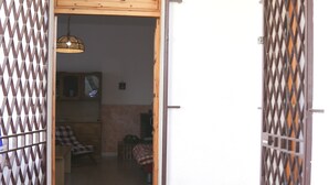 Une porte en bois ouverte donne sur une pièce dont le sol est recouvert de carreaux et où l’on aperçoit un couvre-lit à carreaux.