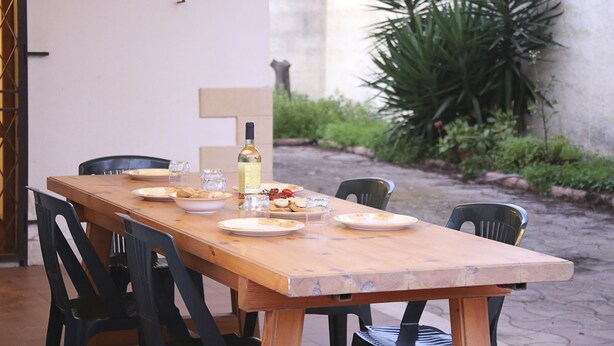 Une table de salle à manger en bois, mise avec des assiettes, des verres et une bouteille, entourée de chaises dans un cadre extérieur agrémenté de plantes et d’un mur en pierre.