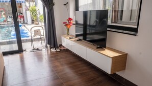A modern living room with a flat-screen TV on a wooden console, a small table with a vase of flowers, and a view of a swimming pool through a glass door. - T&H Hoi An Beach Villa (Da Nang)