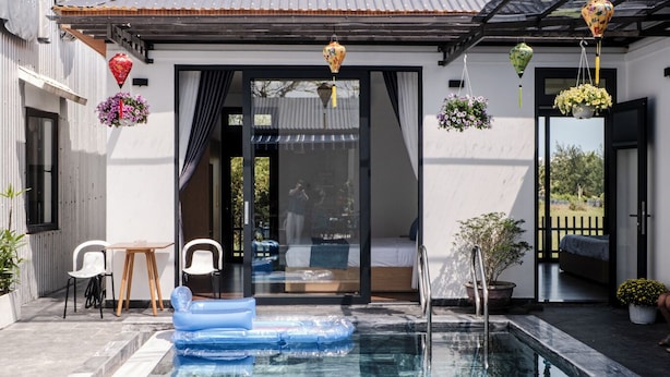 A swimming pool with a clear blue water surface, a wooden deck, and a modern house with a metal roof. - T&H Hoi An Beach Villa (Da Nang)