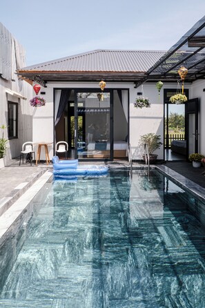 A swimming pool with a clear blue water surface, a wooden deck, and a modern house with a metal roof. - T&H Hoi An Beach Villa (Da Nang)