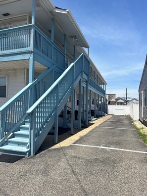A building with a blue staircase leading to a balcony, a parking lot in front, and a clear sky. - Cozy 1-bedroom condo sleeps 2 in peaceful Seaside Heights with parking (Seaside Heights)