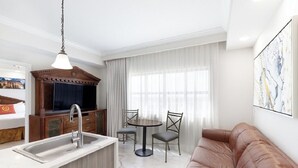 Ein modernes Wohnzimmer mit braunem Ledersofa, einem Essbereich mit rundem Tisch und Stühlen, einem integrierten Fernseher und einer Küche mit Spüle und Holzschränken.