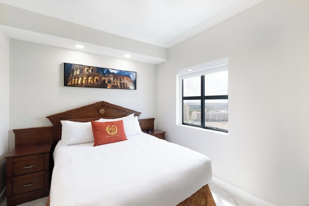 Camera d'albergo con un letto grande, una testiera in legno, un comodino, un cassetto, una finestra con vista e un quadro di un edificio storico appeso al muro.
