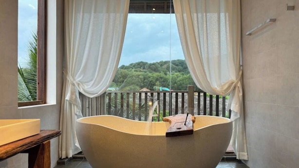 Eine große, freistehende Badewanne in einem Raum mit Blick auf eine üppige Landschaft, die durch transparente Vorhänge zu sehen ist.