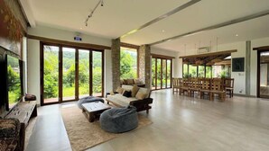 Ein geräumiger Wohnbereich mit einer großen Couch, einem Couchtisch und einem Sitzsack. Es gibt einen Essbereich mit Holztischen und -stühlen. Das Zimmer verfügt über große Fenster mit Blick auf üppiges Grün draußen.