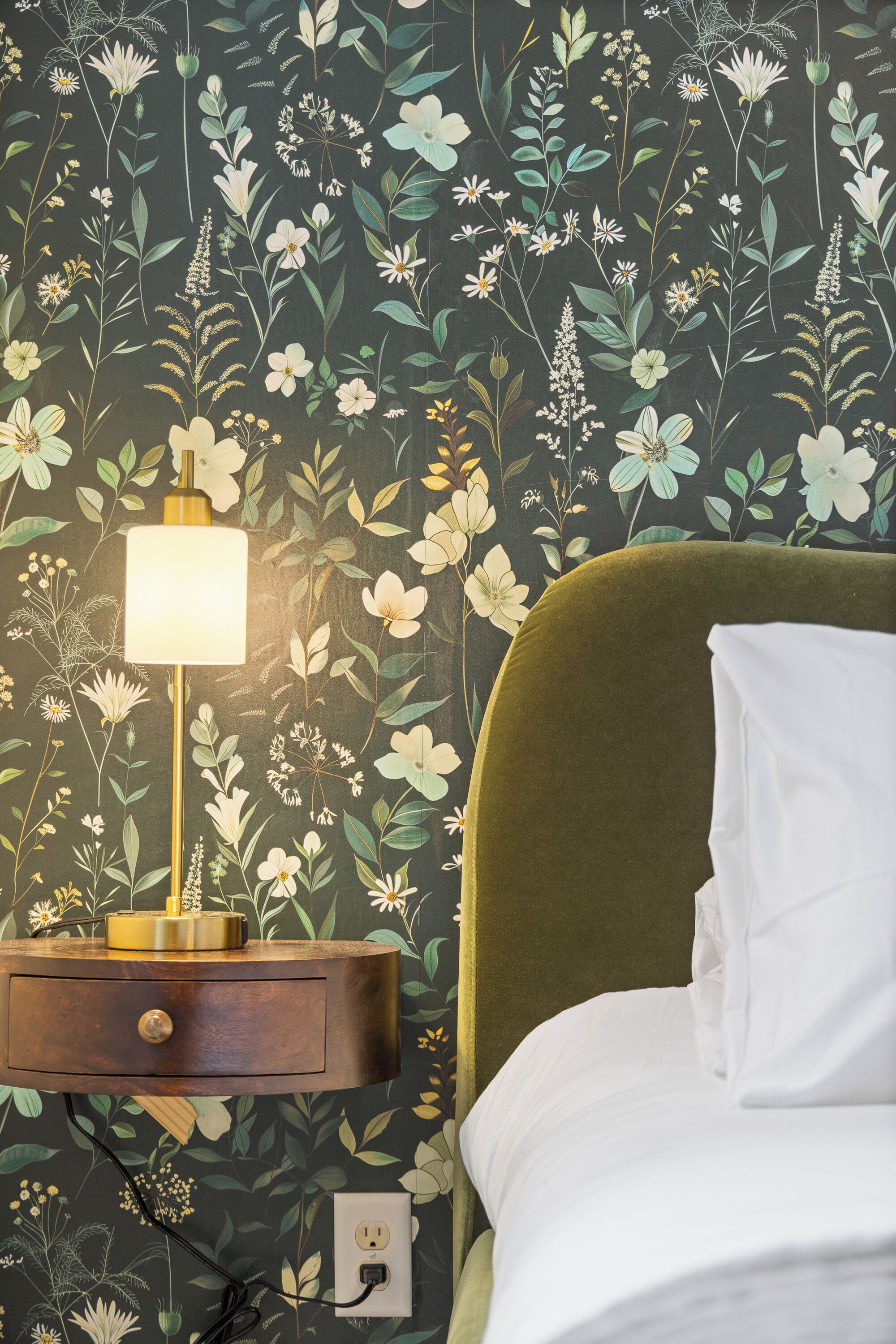 A bedroom with a floral wallpaper, a bedside lamp, a green upholstered headboard, and a white pillow.