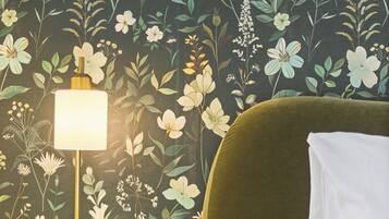 A bedroom with a floral wallpaper, a bedside lamp, a green upholstered headboard, and a white pillow.