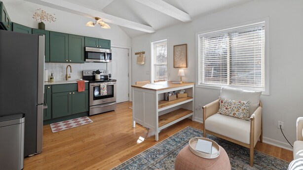 A modern kitchen with green cabinets, a wooden dining table, and a cozy seating area with a chair and ottoman.