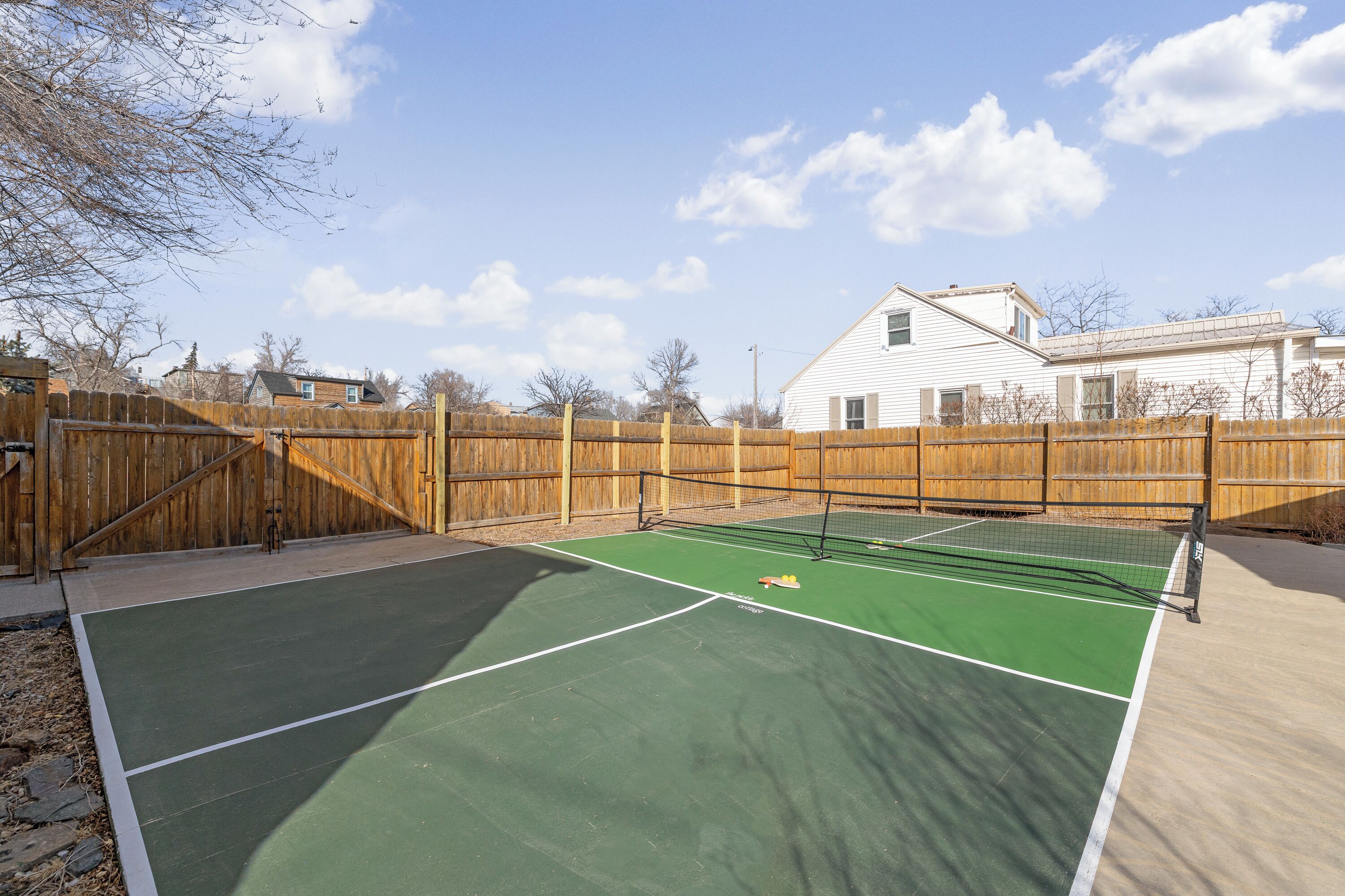Una cancha de tenis con valla de madera, una casa al fondo y cielo despejado.