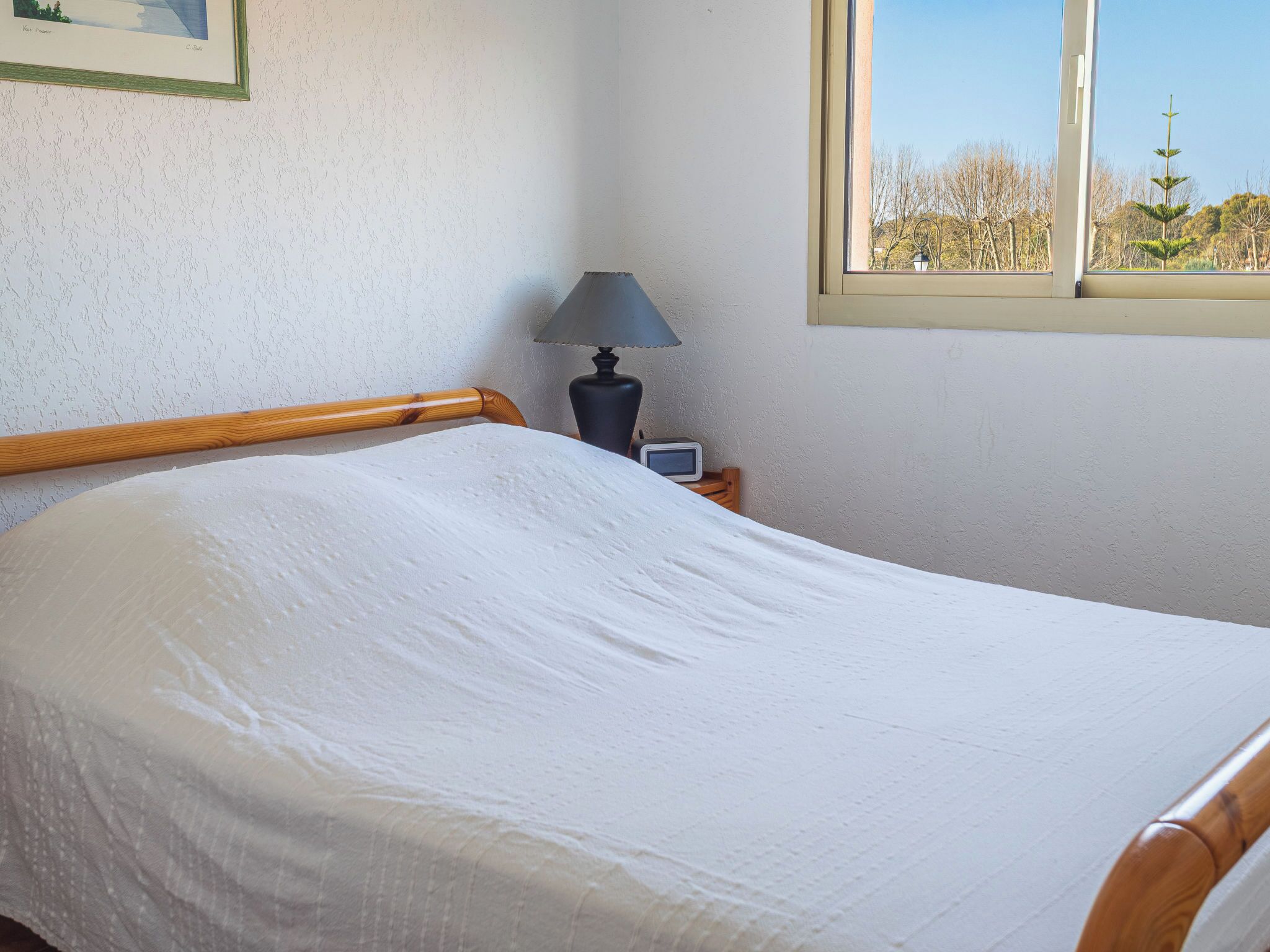 Cama bem arrumada com cabeceira de madeira, luminária de cabeceira e uma mesinha com relógio. Uma janela mostra vista para árvores e um edifício.
