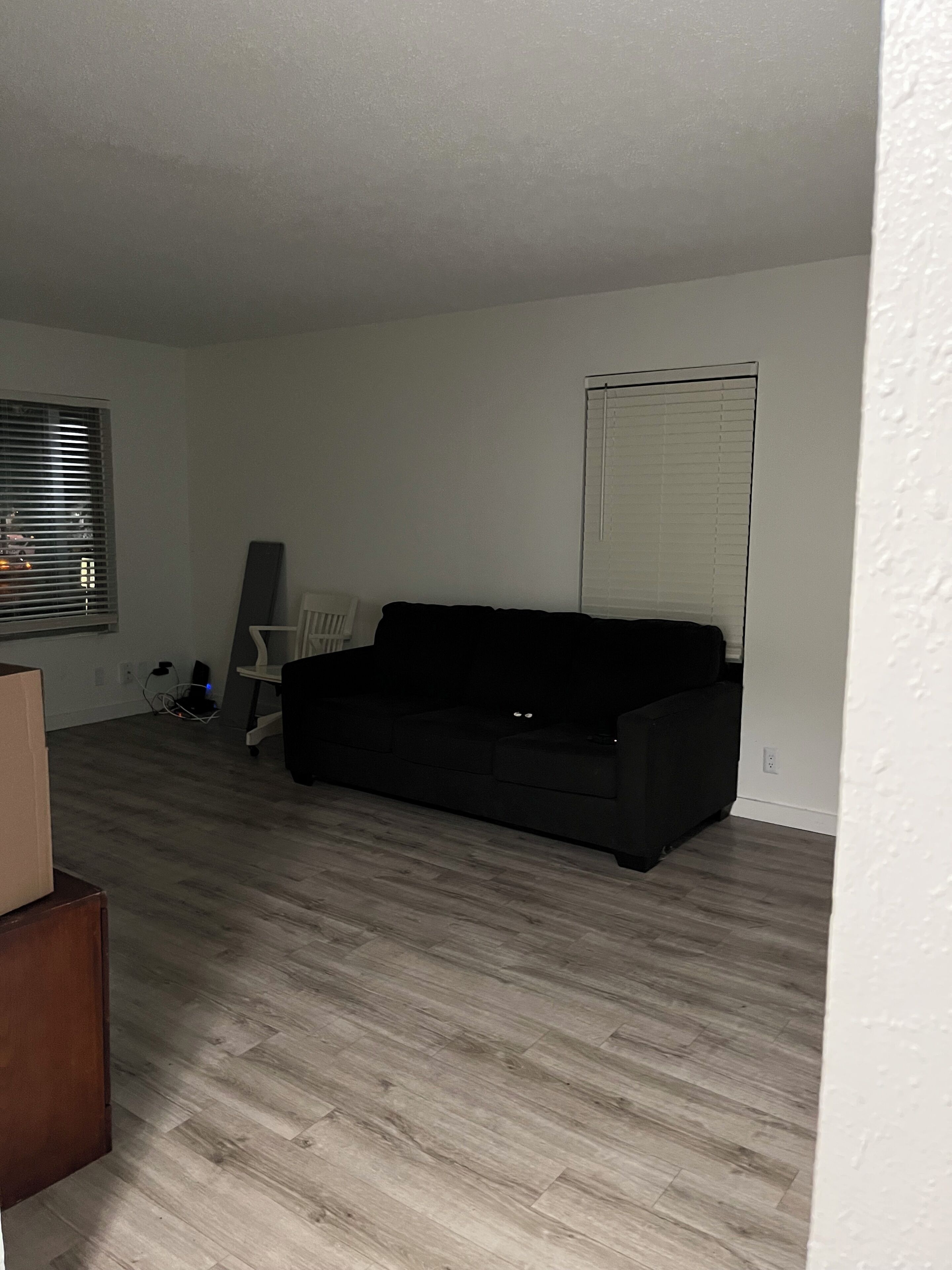 A room with a black sofa, a standing fan, a white chair, and a window with blinds.