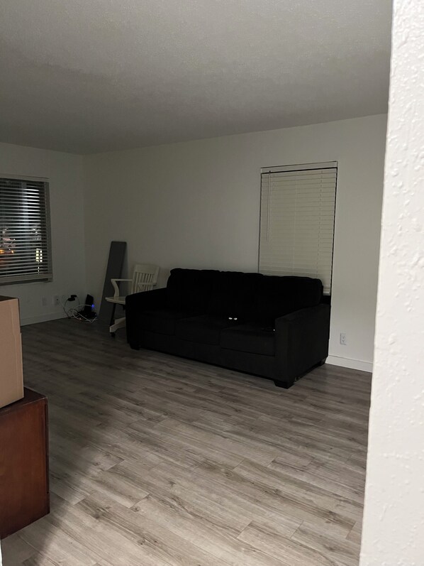 A room with a black sofa, a standing fan, a white chair, and a window with blinds.