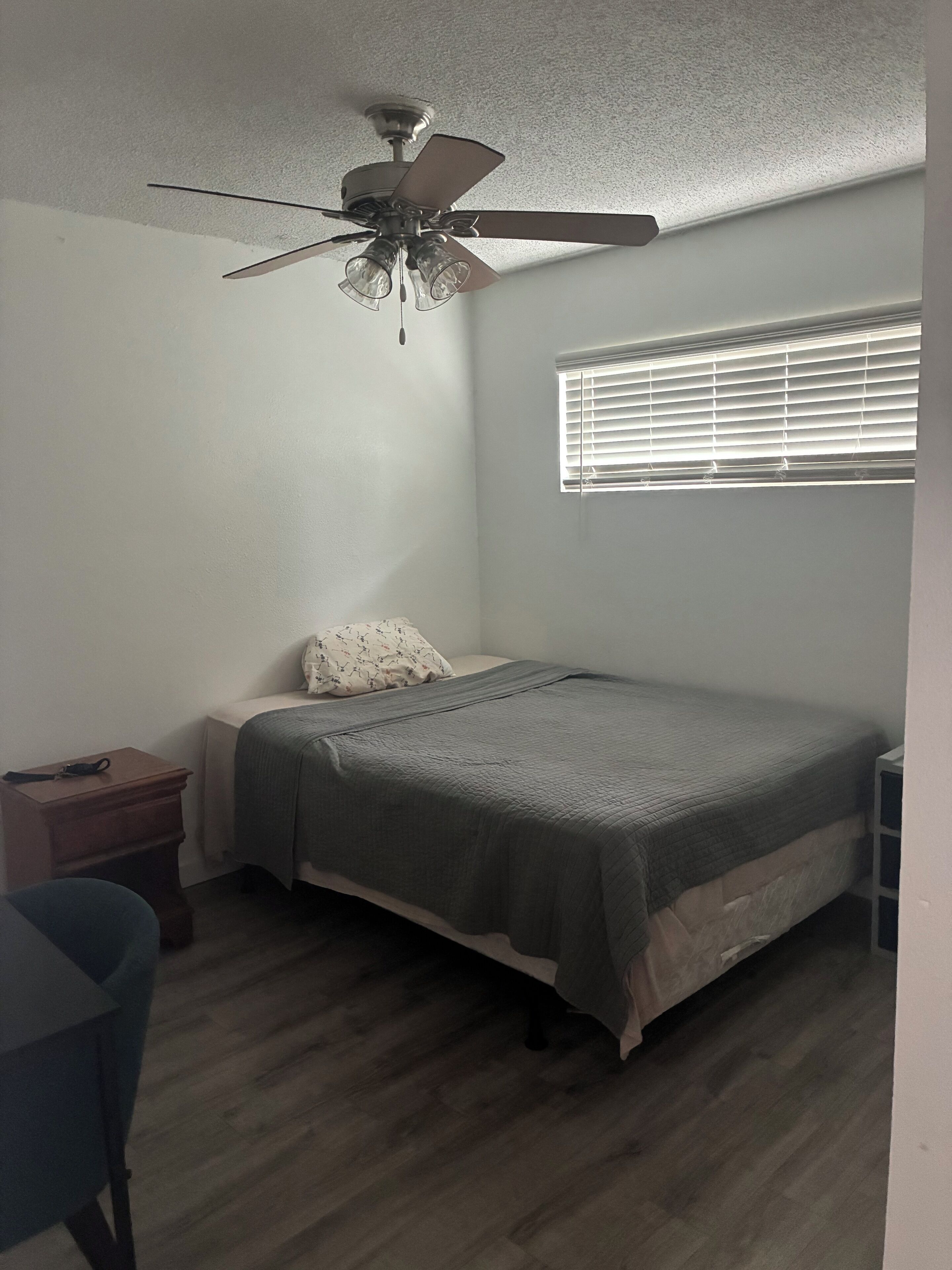 A bedroom with a bed, a ceiling fan, a window with blinds, a nightstand, and a chair.