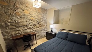Une chambre avec un mur en pierre, un lit avec une literie sombre, un bureau avec une chaise et une table de chevet avec une lampe.