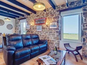 A living room with a stone wall, a leather sofa, a glass coffee table, and a view through a large window. - Vacation home Eden by Interhome (Plouescat)