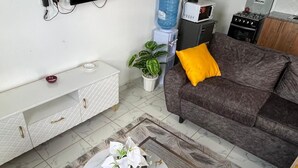 Un salón con un televisor de pantalla plana en la pared, un sofá gris con un cojín amarillo, una mesa de centro blanca con un florero y un control remoto, un armario blanco con cajones, un dispensador de agua y una planta pequeña.