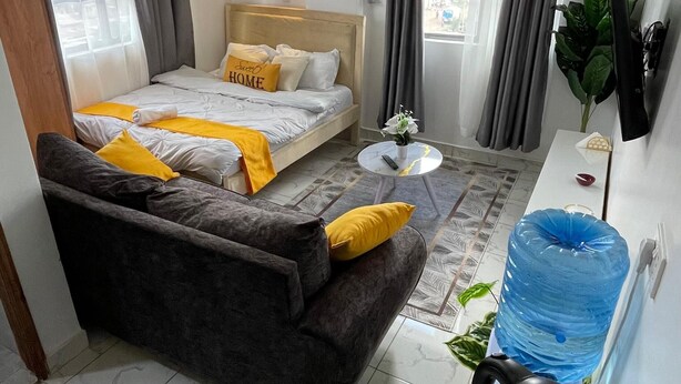 Habitación de hotel compacta con cama, sofá, mesita pequeña, hervidor de agua y dispensador de agua.