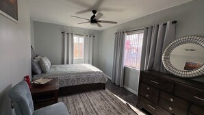 Ein Schlafzimmer mit einem Bett, einem Schminktisch, einem Sessel, einem Deckenventilator und einem Fenster mit Vorhängen.