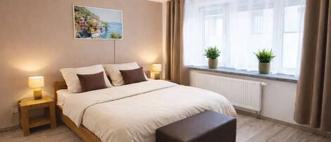 A bedroom with a bed, bedside lamps, a window with curtains, a radiator, and a potted plant.