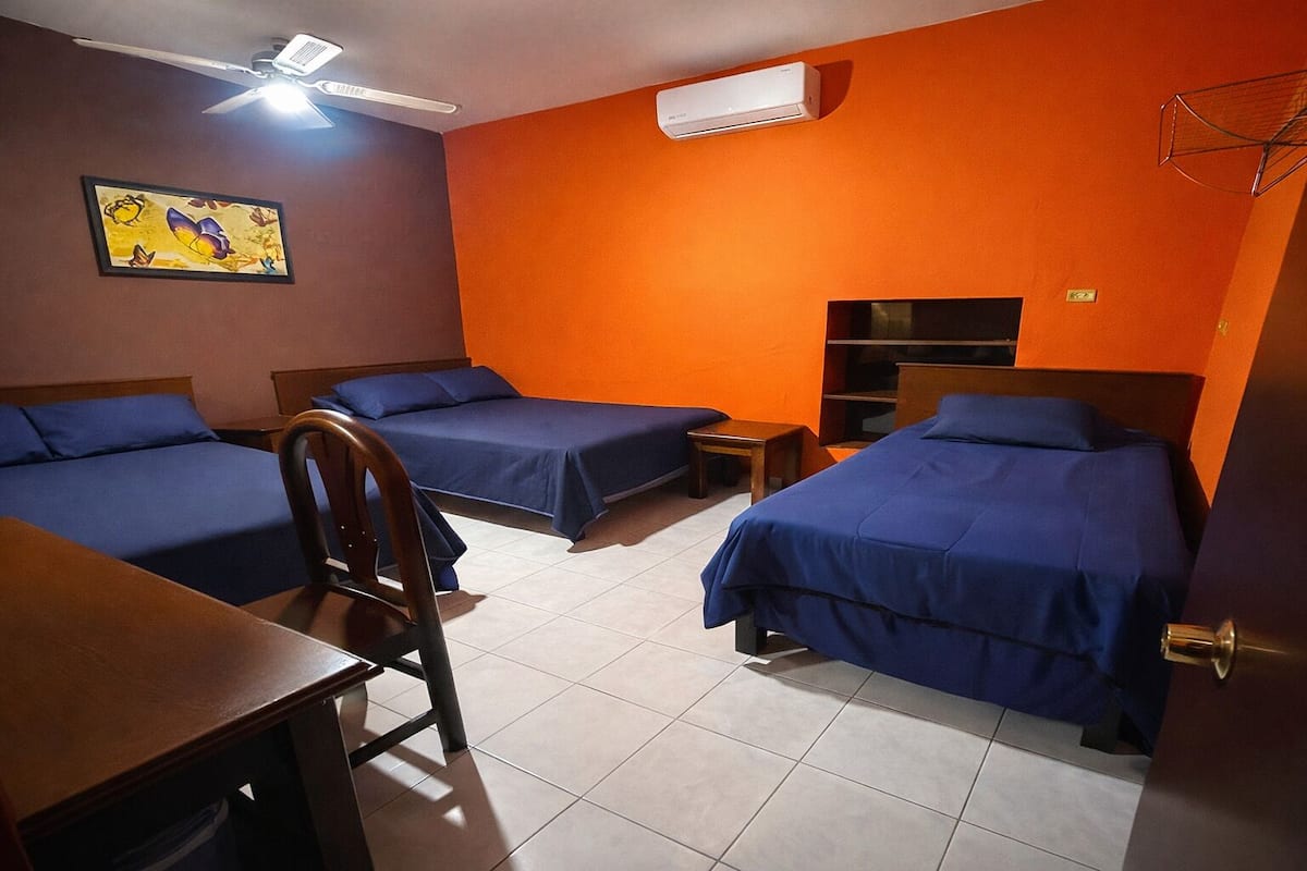 A hotel room with two beds, a desk, a chair, an air conditioner, and a ceiling fan.