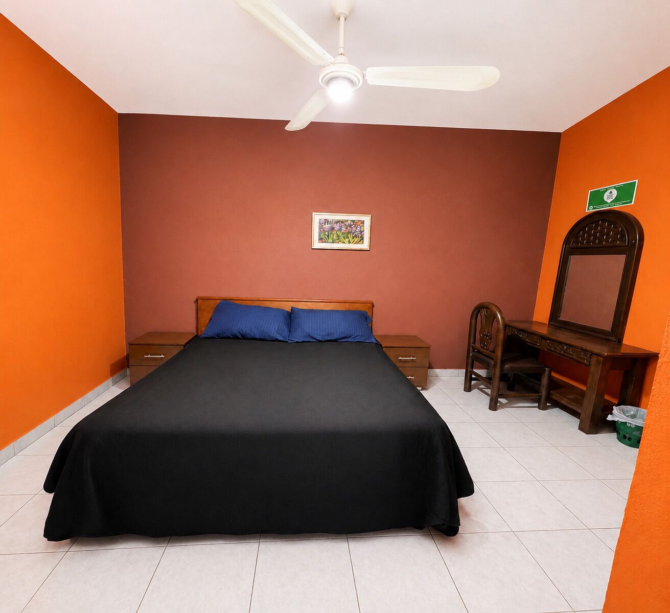 Quarto com cama, escrivaninha, cadeira, ventoinha no teto e um quadro na parede.