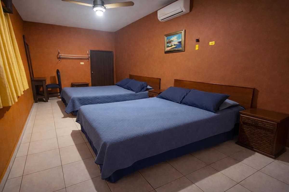 A hotel room with two beds, a ceiling fan, an air conditioner, and a small table with a chair.