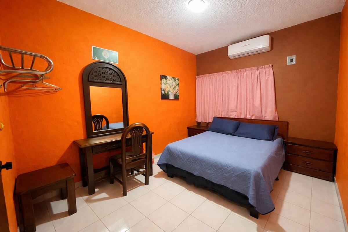 A bedroom with an orange wall, a wooden desk, a chair, a bed with a blue bedspread, a nightstand, a mirror, a window with pink curtains, and an air conditioning unit.