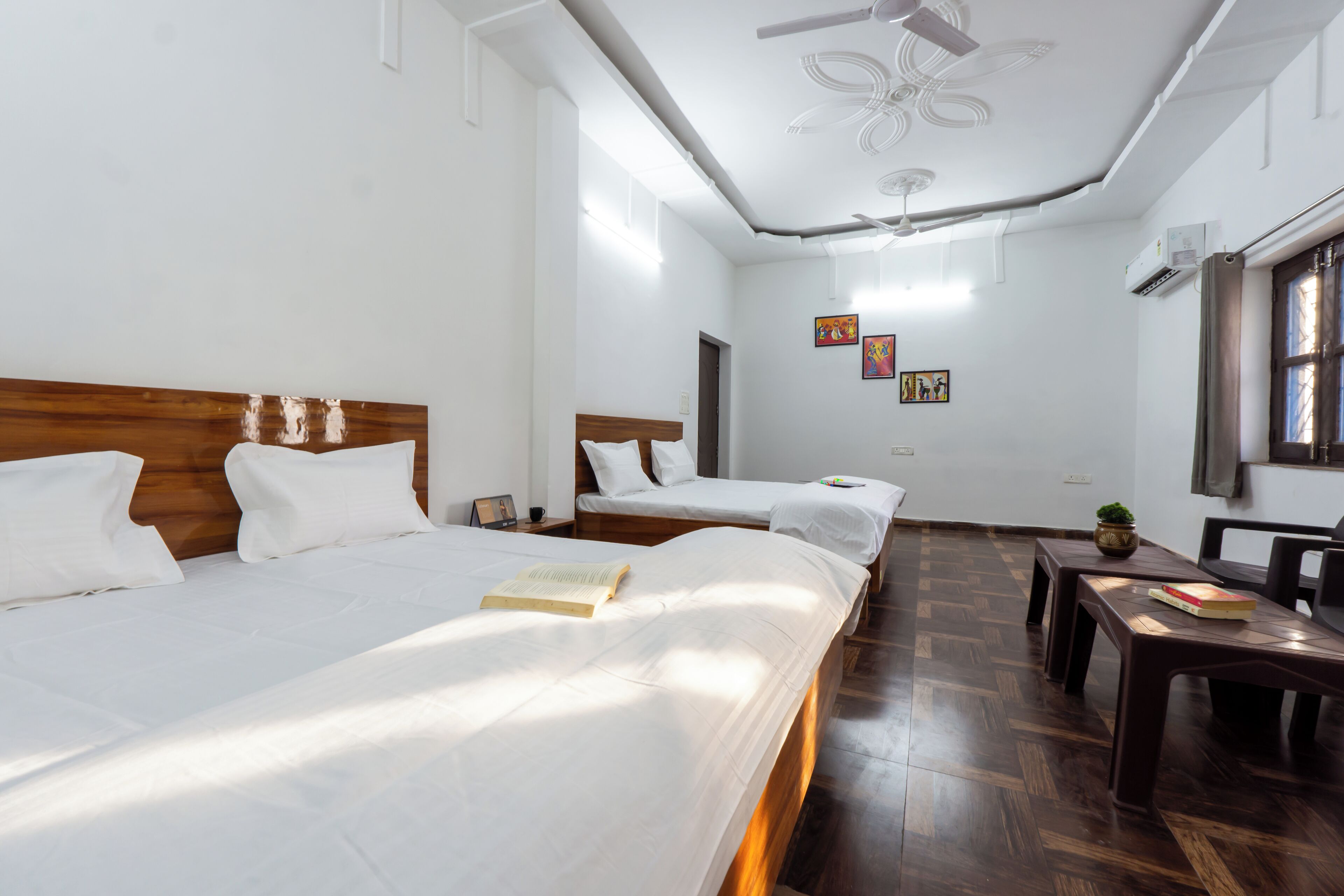 A hotel room with two single beds, a wooden headboard, a small table with a book, and a ceiling fan.