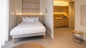 A modern hotel room with a large bed, a small side table, and a kitchenette in the background. - Le Pic Coliving (Clermont-Ferrand)