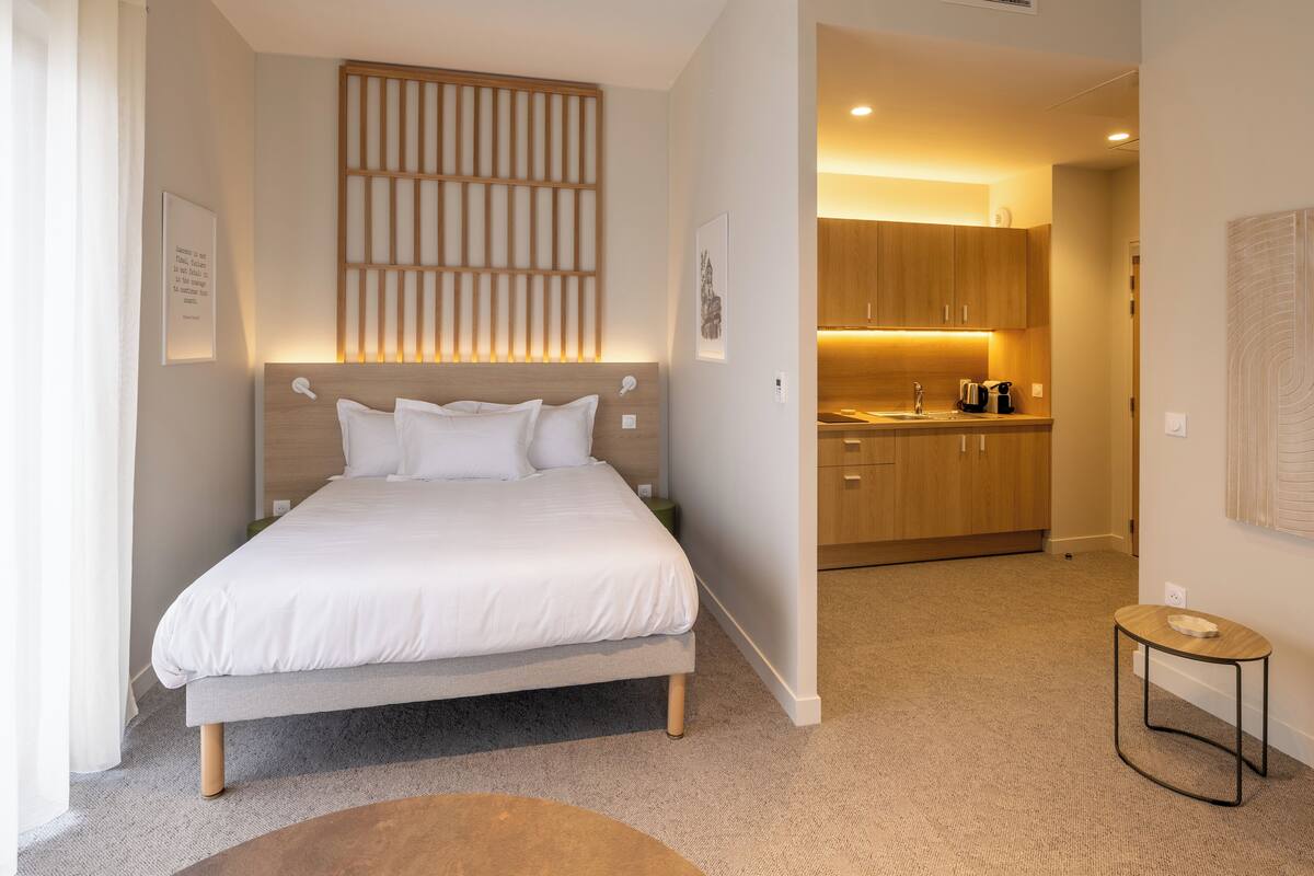 Una habitación de hotel moderna con una cama grande, una mesita auxiliar pequeña y una kitchenette al fondo.