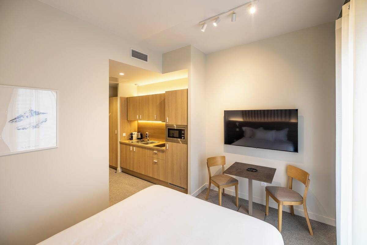Habitación de hotel con cama, una kitchenette pequeña, una zona de comedor con dos sillas y un televisor montado en la pared.