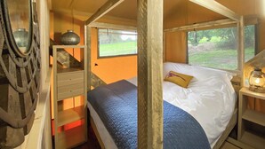A wooden four-poster bed with a white and blue quilt, a small bedside table with a lamp, and a window with a view of a grassy field.