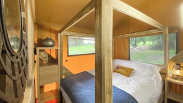 A wooden four-poster bed with a white and blue quilt, a small bedside table with a lamp, and a window with a view of a grassy field.