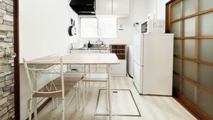 Een compacte keuken met witte kastjes, een magnetron en een fornuis. Er is een eettafel met stoelen en een raam boven de gootsteen.