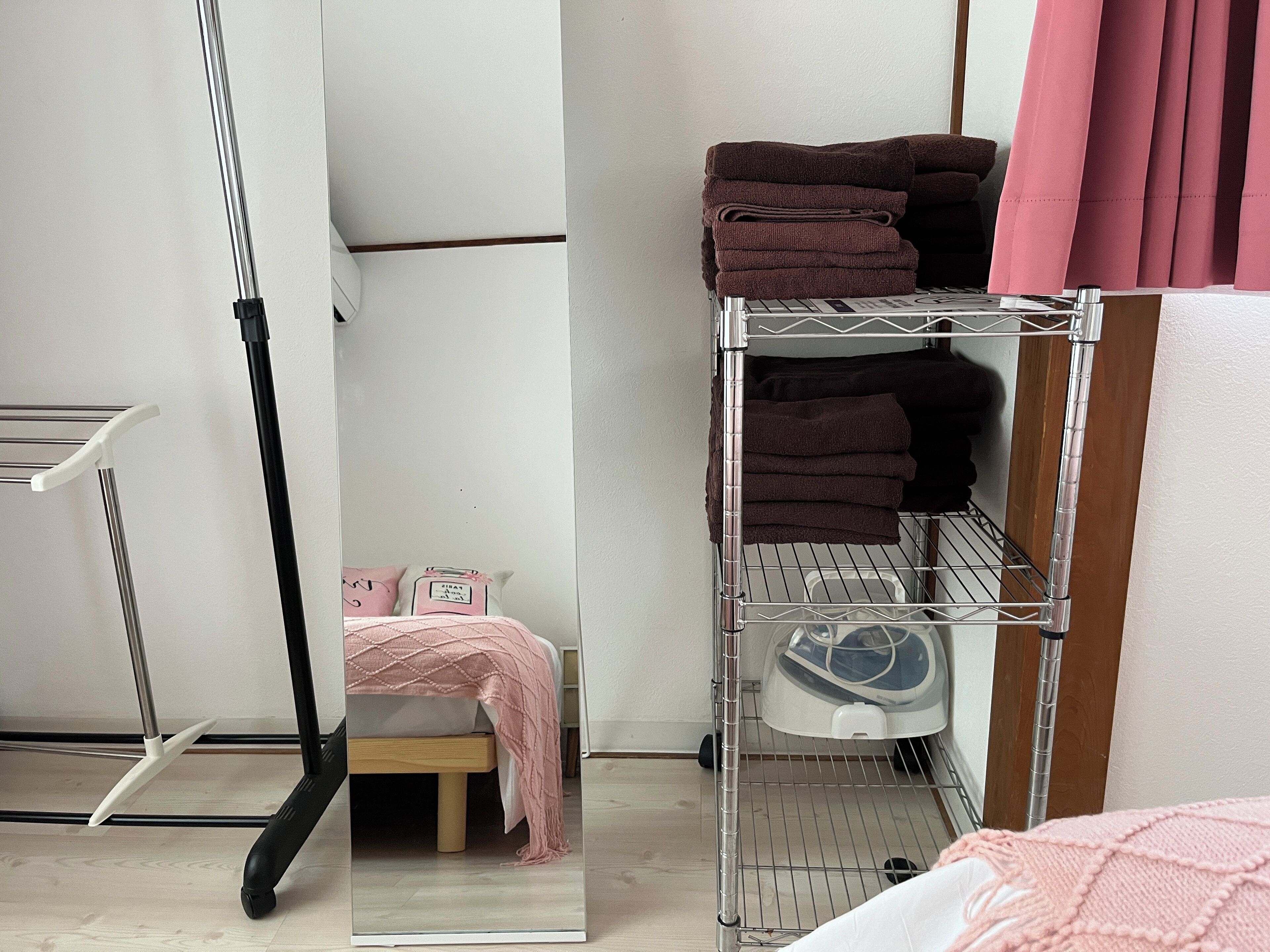 A compact room with a metal shelf unit for storage, a small bedside table with a pink blanket, and a mirror reflecting the room's interior.