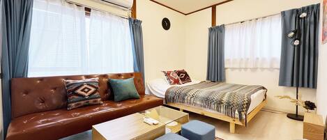 Habitación de hotel compacta con un sofá de cuero marrón, una mesa de centro de madera, dos otomanas azules, una cama de madera con ropa de cama estampada y una alfombra roja y blanca.