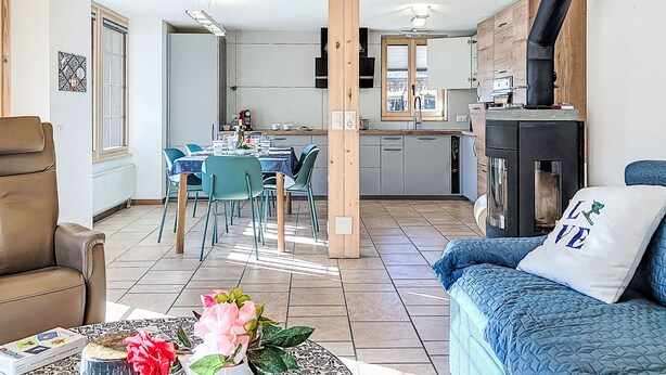 Uma cozinha moderna com fogão embutido, sala de jantar com mesa e cadeiras, e sala de estar com sofá e almofada decorativa.